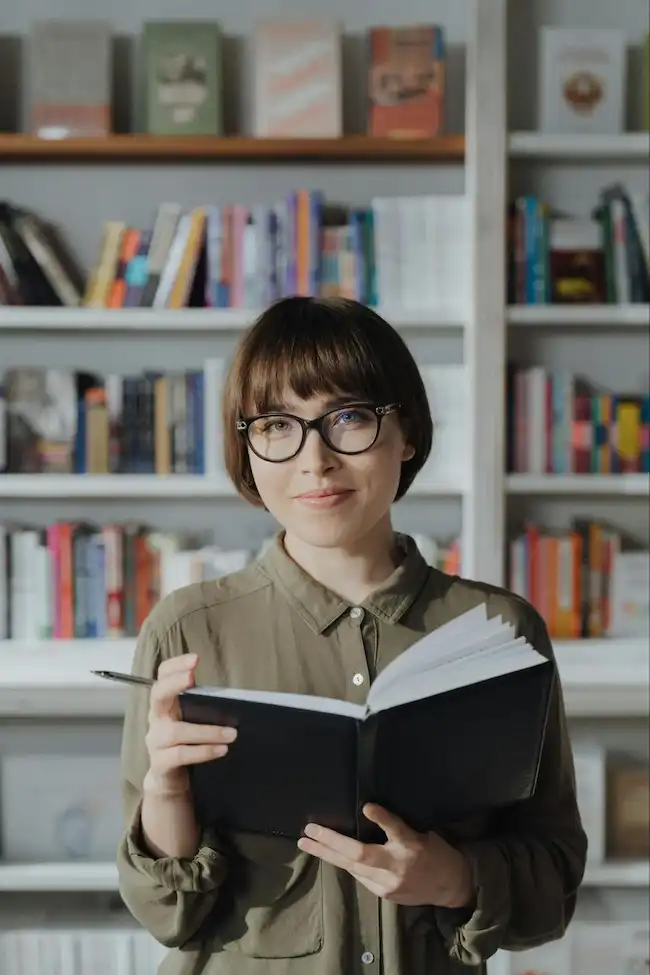 Mujer joven con lentes, sosteniendo un libro abierto en una biblioteca, mirando a la cámara con una expresión tranquila.